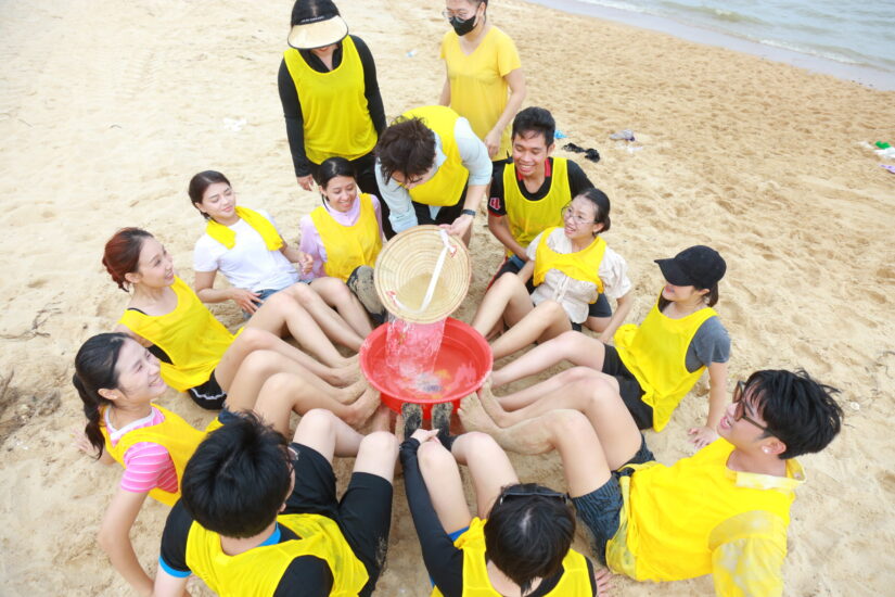 Thành viên áo vàng quây tròn chơi trò chơi đồng đội trên bãi cát