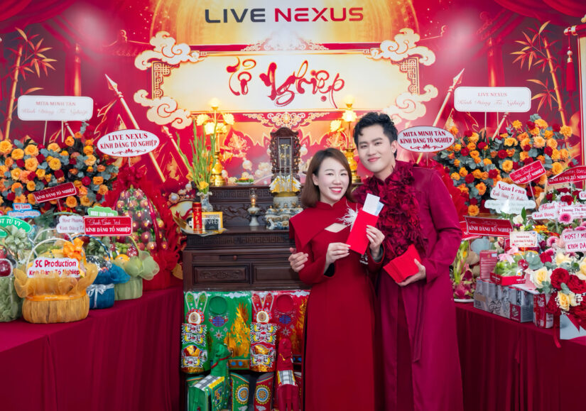 Hai thành viên mặc áo dài đỏ đứng cạnh bàn thờ Tổ nghiệp dưới bảng hiệu LIVE NEXUS